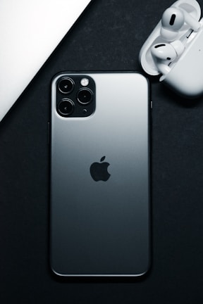 A silver smartphone with the Apple logo prominently displayed is placed against a dark background. Next to it, white wireless earbuds are partially visible in their open charging case.