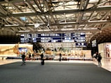 A busy airport terminal with a large decorative sculpture featuring mythical figures. Overhead signs display various gate numbers, and travelers are moving around with luggage. The area is well-lit with modern architectural elements visible in the ceiling structure.