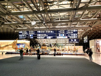 A busy airport terminal with a large decorative sculpture featuring mythical figures. Overhead signs display various gate numbers, and travelers are moving around with luggage. The area is well-lit with modern architectural elements visible in the ceiling structure.