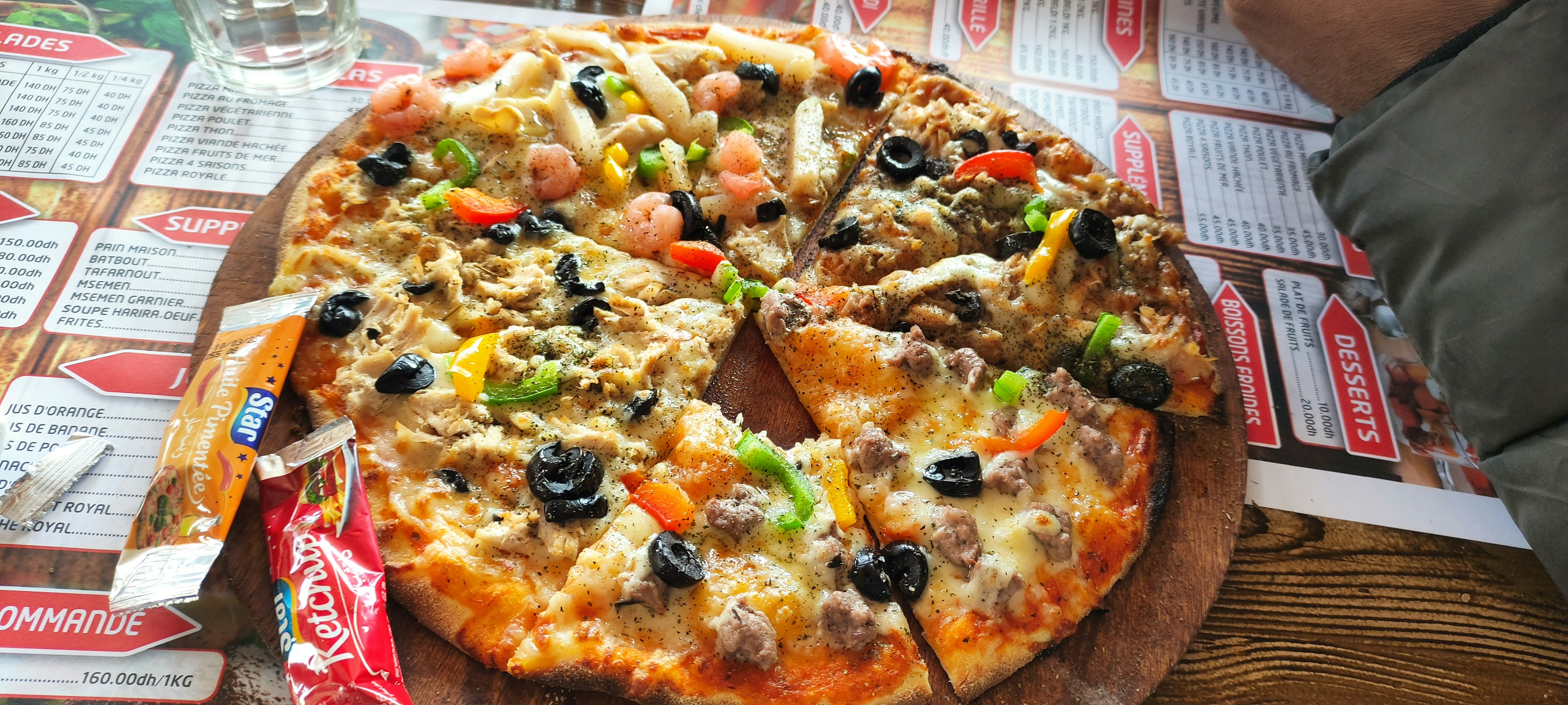 A pizza topped with a variety of ingredients including olives, bell peppers, shrimp, and cheese is placed on a wooden serving board. Surrounding the pizza are condiment packets and a glass of water. The table is covered with a menu featuring various dishes.