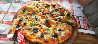 A pizza topped with a variety of ingredients including olives, bell peppers, shrimp, and cheese is placed on a wooden serving board. Surrounding the pizza are condiment packets and a glass of water. The table is covered with a menu featuring various dishes.