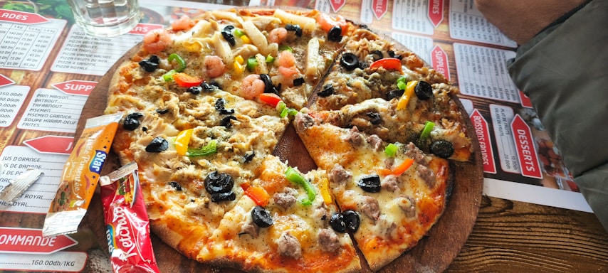 A pizza topped with a variety of ingredients including olives, bell peppers, shrimp, and cheese is placed on a wooden serving board. Surrounding the pizza are condiment packets and a glass of water. The table is covered with a menu featuring various dishes.