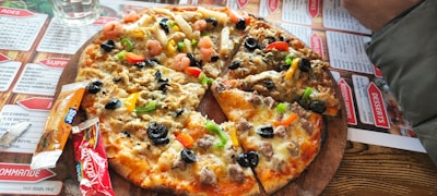 A pizza topped with a variety of ingredients including olives, bell peppers, shrimp, and cheese is placed on a wooden serving board. Surrounding the pizza are condiment packets and a glass of water. The table is covered with a menu featuring various dishes.
