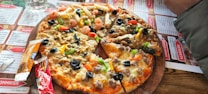 A pizza topped with a variety of ingredients including olives, bell peppers, shrimp, and cheese is placed on a wooden serving board. Surrounding the pizza are condiment packets and a glass of water. The table is covered with a menu featuring various dishes.