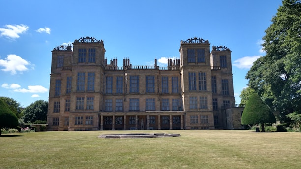 A grand, historic mansion with multiple tall windows and intricate architectural details. It stands on a well-kept lawn with symmetrical topiary trees on either side. The building is set against a bright blue sky with a few scattered clouds.