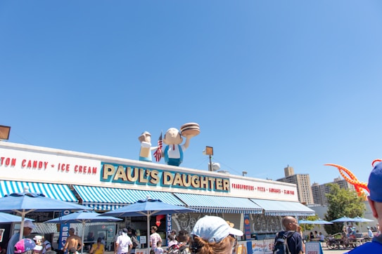 A vibrant outdoor scene featuring a classic seaside eatery with a marquee that reads 'Paul's Daughter'. The storefront is adorned with blue and white stripes, and there are several people nearby, some of whom are waiting in line. Oversized cartoonish figures hold a hotdog and ice cream cone on the roof, and a small American flag is visible. The sky is clear and blue, adding to the cheerful atmosphere.