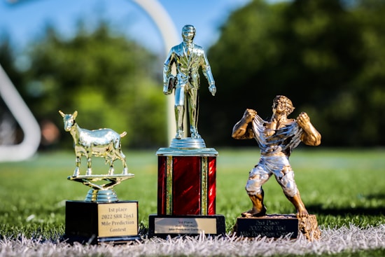 Three trophies are displayed on a grassy surface under bright sunlight. The trophy on the left features a goat on a black base with an inscription reading '1st place 2022 SRR 26x1 Mile Prediction Contest'. The middle trophy is a classic, golden figurine of a man in a suit holding a briefcase, standing on a red pedestal base with an inscription for '3rd Place'. The trophy on the right depicts a muscular man ripping his shirt, standing on a black base labeled '2nd Place 2022 SRR 26x1 Mile Prediction Contest'.