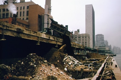 An industrial area by the waterfront features a large pile of rubble and debris. A truck is positioned at the edge of a structure, unloading more materials onto the heap. In the background, smokestacks and tall buildings rise into a gray, overcast sky, suggesting an urban environment.