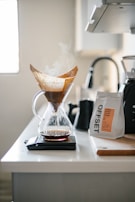 A kitchen counter displays a glass pour-over coffee maker with steam rising from freshly brewed coffee. The filter with coffee grounds is visible, and the decanter sits atop a digital scale. Nearby, a bag of coffee beans labeled 'OFFSET' is placed beside a black kettle and a knife block.