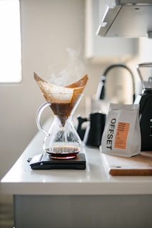 A kitchen counter displays a glass pour-over coffee maker with steam rising from freshly brewed coffee. The filter with coffee grounds is visible, and the decanter sits atop a digital scale. Nearby, a bag of coffee beans labeled 'OFFSET' is placed beside a black kettle and a knife block.