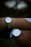 Two wrists are closely positioned, each wearing a wristwatch. The watches have a matching design and feature a silver and gold metal band with a classic white watch face and minimalistic details.