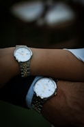 Two wrists are closely positioned, each wearing a wristwatch. The watches have a matching design and feature a silver and gold metal band with a classic white watch face and minimalistic details.