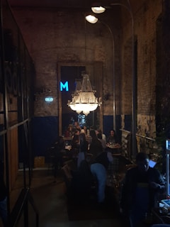 A dimly lit, industrial-style interior of a restaurant or bar with exposed brick walls and a large chandelier overhead. People are seated at tables, engaging in conversation and dining. The atmosphere is cozy and intimate with a mix of modern and rustic elements.