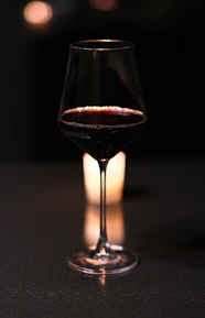 A glass of deep red wine catching warm candlelight on a matte black table.