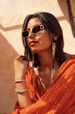 A person with short dark hair wearing stylish sunglasses, large looped gold earrings, and a vibrant orange outfit. The background has a warm, sunlit tone, contributing to a chic and confident vibe. The person is accessorized with bracelets and a fashion chain that accenuates the ensemble.