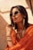 A person with short dark hair wearing stylish sunglasses, large looped gold earrings, and a vibrant orange outfit. The background has a warm, sunlit tone, contributing to a chic and confident vibe. The person is accessorized with bracelets and a fashion chain that accenuates the ensemble.