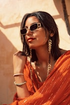 A person with short dark hair wearing stylish sunglasses, large looped gold earrings, and a vibrant orange outfit. The background has a warm, sunlit tone, contributing to a chic and confident vibe. The person is accessorized with bracelets and a fashion chain that accenuates the ensemble.