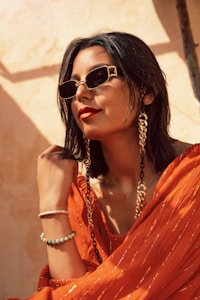 A person with short dark hair wearing stylish sunglasses, large looped gold earrings, and a vibrant orange outfit. The background has a warm, sunlit tone, contributing to a chic and confident vibe. The person is accessorized with bracelets and a fashion chain that accenuates the ensemble.