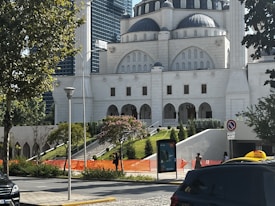 A large, white building with traditional Islamic architectural elements, featuring domes and arched windows. Trees and greenery surround the building, and a few pedestrians are visible near the entrance. Cars, including a taxi, are on the nearby street, along with construction barriers.