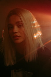 A person with long blonde hair is softly illuminated by warm light, creating a dreamy and ethereal effect. The light reflects off the hair, producing a halo-like glow. The background is dark, emphasizing the contrasting illumination on the subject's face and hair.