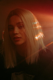A person with long blonde hair is softly illuminated by warm light, creating a dreamy and ethereal effect. The light reflects off the hair, producing a halo-like glow. The background is dark, emphasizing the contrasting illumination on the subject's face and hair.