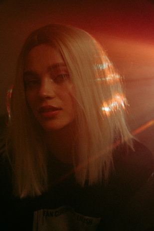 A person with long blonde hair is softly illuminated by warm light, creating a dreamy and ethereal effect. The light reflects off the hair, producing a halo-like glow. The background is dark, emphasizing the contrasting illumination on the subject's face and hair.