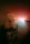 A young woman with blonde hair is captured in a dreamy, ethereal setting. She has her eyes closed and wears a dark t-shirt featuring a graphic print. A bright, colorful light source behind her creates a halo effect, casting soft pink, red, and orange hues across the scene, giving a surreal and tranquil atmosphere.