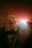 A young woman with blonde hair is captured in a dreamy, ethereal setting. She has her eyes closed and wears a dark t-shirt featuring a graphic print. A bright, colorful light source behind her creates a halo effect, casting soft pink, red, and orange hues across the scene, giving a surreal and tranquil atmosphere.