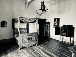 An antique room featuring a large, ornately carved wooden bed with intricate detailing and a canopy. The room includes a vintage wardrobe, a decorative chest of drawers, and a mounted animal head on the wall. A portrait of a historical figure hangs above the chest, adding to the vintage ambiance. The room is illuminated by soft light, creating shadows and highlights on the aged wooden floor.
