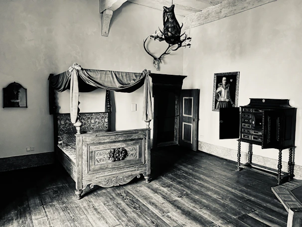 An antique room featuring a large, ornately carved wooden bed with intricate detailing and a canopy. The room includes a vintage wardrobe, a decorative chest of drawers, and a mounted animal head on the wall. A portrait of a historical figure hangs above the chest, adding to the vintage ambiance. The room is illuminated by soft light, creating shadows and highlights on the aged wooden floor.
