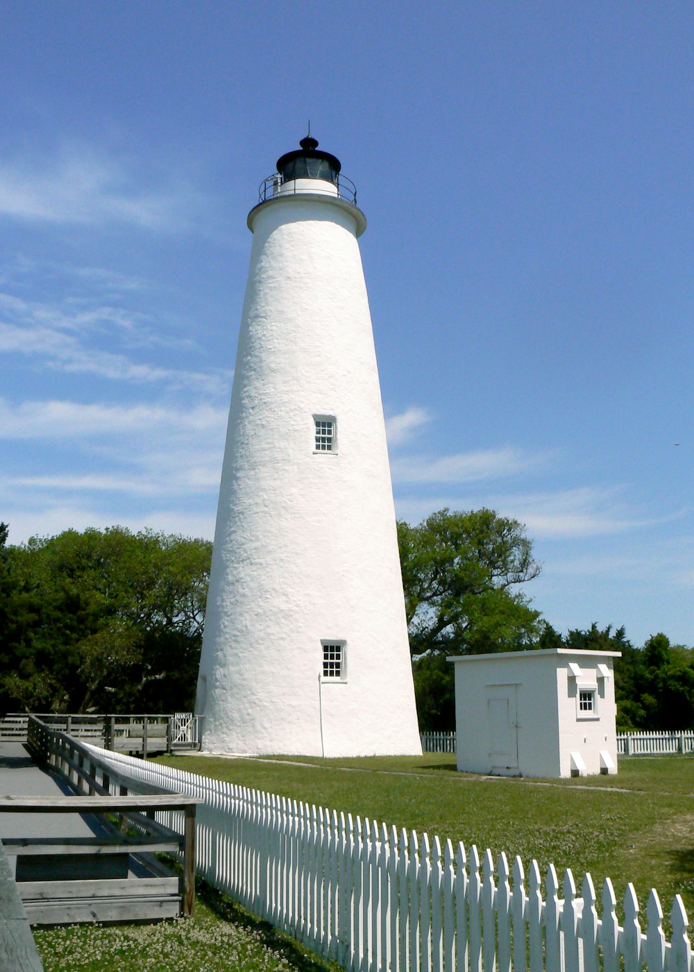 a white lighthouse with a white fence with Ocracoke in the background