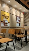 A cozy café interior with wooden chairs and tables neatly arranged. On the wall are two large posters advertising different beverages: one featuring a caramel frappe with the text 'Wintry Vibes', and another showcasing hot drinks with 'Hots for You'. The lighting is warm and the setting is modern with clean lines.