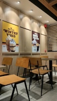 A cozy café interior with wooden chairs and tables neatly arranged. On the wall are two large posters advertising different beverages: one featuring a caramel frappe with the text 'Wintry Vibes', and another showcasing hot drinks with 'Hots for You'. The lighting is warm and the setting is modern with clean lines.