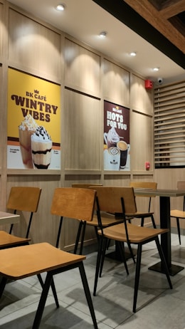 A cozy café interior with wooden chairs and tables neatly arranged. On the wall are two large posters advertising different beverages: one featuring a caramel frappe with the text 'Wintry Vibes', and another showcasing hot drinks with 'Hots for You'. The lighting is warm and the setting is modern with clean lines.