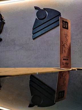 A decorative wall display with a modern, stylized black silhouette of a slice of cake with a cherry on top. Next to it, a vertical wooden block with lettering that says 'Home of #Desserts' along with additional text and the number 01. The setup is reflected on a glossy surface beneath.