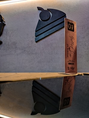 A decorative wall display with a modern, stylized black silhouette of a slice of cake with a cherry on top. Next to it, a vertical wooden block with lettering that says 'Home of #Desserts' along with additional text and the number 01. The setup is reflected on a glossy surface beneath.
