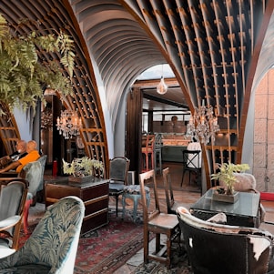 An eclectic interior space featuring ornate wooden architectural elements forming arches overhead. The room is furnished with a mix of vintage and modern chairs and tables. Crystal chandeliers hang from the ceiling, adding a touch of elegance. Various potted plants are placed around, contributing to a cozy ambiance. The decor includes rich textiles and intricate patterns, creating an inviting atmosphere.