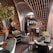 An eclectic interior space featuring ornate wooden architectural elements forming arches overhead. The room is furnished with a mix of vintage and modern chairs and tables. Crystal chandeliers hang from the ceiling, adding a touch of elegance. Various potted plants are placed around, contributing to a cozy ambiance. The decor includes rich textiles and intricate patterns, creating an inviting atmosphere.