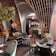 An eclectic interior space featuring ornate wooden architectural elements forming arches overhead. The room is furnished with a mix of vintage and modern chairs and tables. Crystal chandeliers hang from the ceiling, adding a touch of elegance. Various potted plants are placed around, contributing to a cozy ambiance. The decor includes rich textiles and intricate patterns, creating an inviting atmosphere.