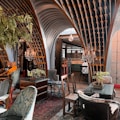 An eclectic interior space featuring ornate wooden architectural elements forming arches overhead. The room is furnished with a mix of vintage and modern chairs and tables. Crystal chandeliers hang from the ceiling, adding a touch of elegance. Various potted plants are placed around, contributing to a cozy ambiance. The decor includes rich textiles and intricate patterns, creating an inviting atmosphere.