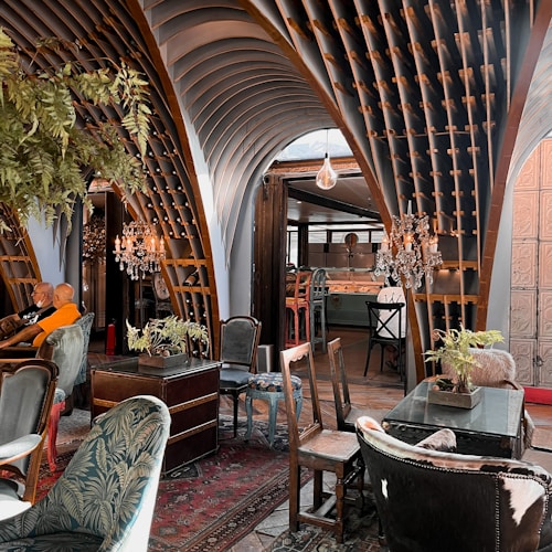 An eclectic interior space featuring ornate wooden architectural elements forming arches overhead. The room is furnished with a mix of vintage and modern chairs and tables. Crystal chandeliers hang from the ceiling, adding a touch of elegance. Various potted plants are placed around, contributing to a cozy ambiance. The decor includes rich textiles and intricate patterns, creating an inviting atmosphere.