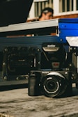 A Sony camera equipped with a Godox flash is placed on a wooden surface. In the background, a blurred figure can be seen, suggesting a person standing behind some metal cases or equipment.