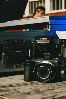 A Sony camera equipped with a Godox flash is placed on a wooden surface. In the background, a blurred figure can be seen, suggesting a person standing behind some metal cases or equipment.