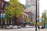 A city street with a row of parked cars along the side, lined with brick buildings and street signs. Trees with fresh green leaves are visible, and a pedestrian is waiting at a crosswalk. The street is marked by a traffic light displaying green, and a sign that reads 'Robson St' is present.