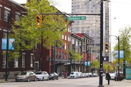 A city street with a row of parked cars along the side, lined with brick buildings and street signs. Trees with fresh green leaves are visible, and a pedestrian is waiting at a crosswalk. The street is marked by a traffic light displaying green, and a sign that reads 'Robson St' is present.