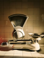 A vintage weighing scale sits against a tiled wall, with a dramatic play of light and shadow highlighting its metallic and ceramic surfaces. The scale has a large dial marked 'Mercado McCann' and a pan for weighing items. The tiles are arranged in a neat, grid-like pattern, contributing to a timeless atmosphere.