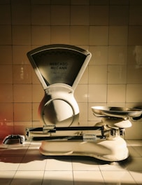 A vintage weighing scale sits against a tiled wall, with a dramatic play of light and shadow highlighting its metallic and ceramic surfaces. The scale has a large dial marked 'Mercado McCann' and a pan for weighing items. The tiles are arranged in a neat, grid-like pattern, contributing to a timeless atmosphere.