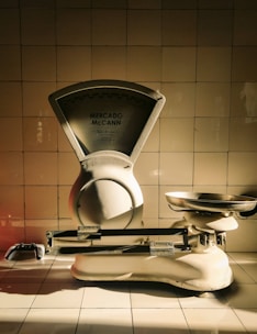 A vintage weighing scale sits against a tiled wall, with a dramatic play of light and shadow highlighting its metallic and ceramic surfaces. The scale has a large dial marked 'Mercado McCann' and a pan for weighing items. The tiles are arranged in a neat, grid-like pattern, contributing to a timeless atmosphere.