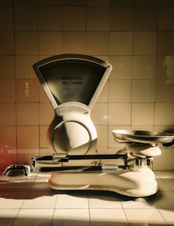 A vintage weighing scale sits against a tiled wall, with a dramatic play of light and shadow highlighting its metallic and ceramic surfaces. The scale has a large dial marked 'Mercado McCann' and a pan for weighing items. The tiles are arranged in a neat, grid-like pattern, contributing to a timeless atmosphere.