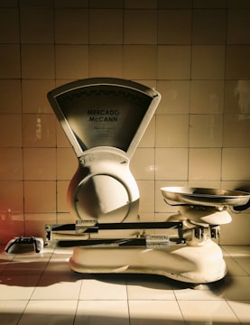 A vintage weighing scale sits against a tiled wall, with a dramatic play of light and shadow highlighting its metallic and ceramic surfaces. The scale has a large dial marked 'Mercado McCann' and a pan for weighing items. The tiles are arranged in a neat, grid-like pattern, contributing to a timeless atmosphere.